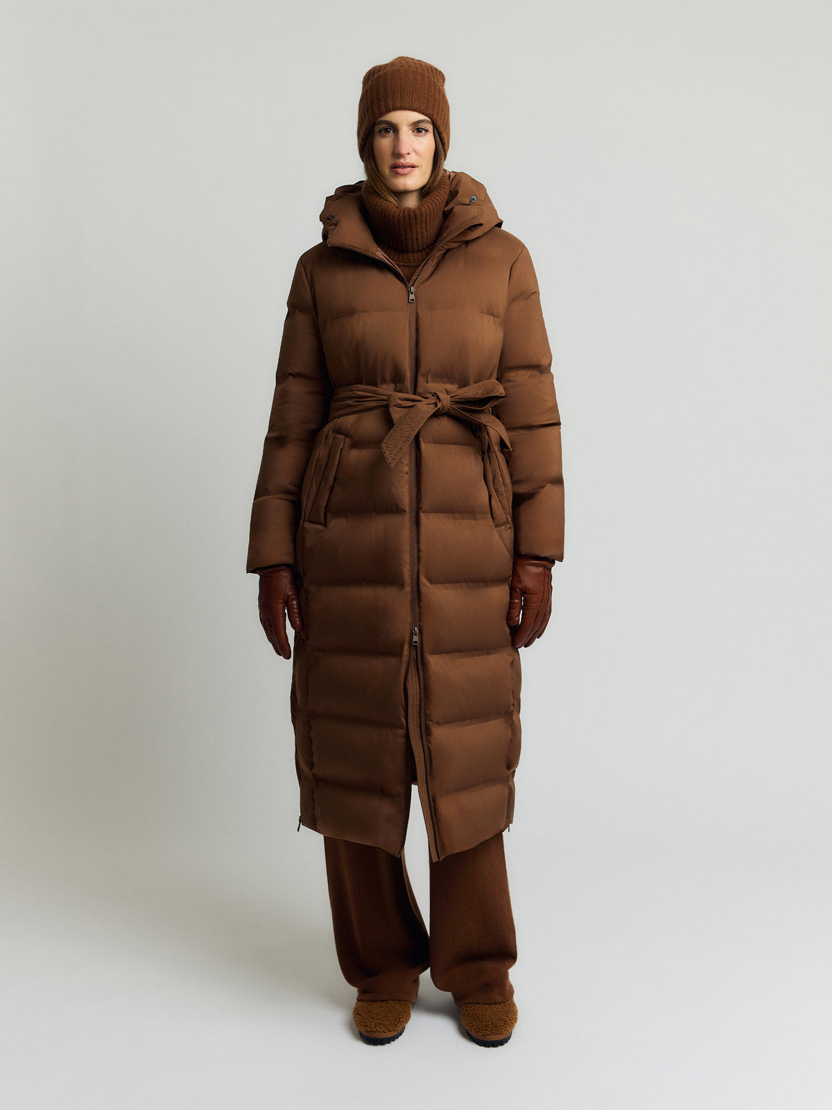 Pitkä ruskea untuvatakki hupulla, kaksisuuntainen vetoketju, solmittava vyö ja vaakatikkaus päällä edestä